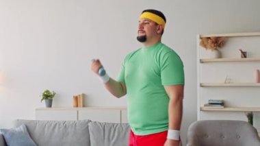 Bin yıllık bir adam evinde spor yapıyor, spor yapmaya ve kilo vermeye odaklanıyor. Rahat bir spor kıyafeti giyer ve iyi dekore edilmiş bir salonda ağırlık kullanır..