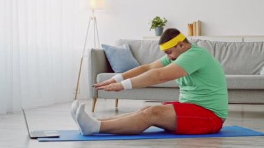 Rahat idman kıyafeti giyen bir adam oturma odasındaki paspasın üzerine uzanır. Kilo vermek için bir online egzersiz programını takip ederken formda kalmaya odaklanıyor..