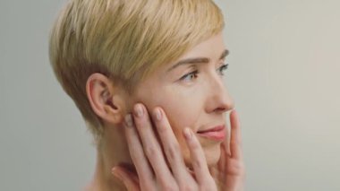 50 'li yaşlarında beyaz bir kadın yüzüne cilt ürünleri sürüyor. Kısa saçı, kendini şımartmaya odaklanırken rahat yüzünü çerçeveler..