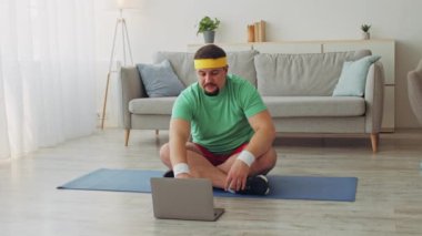 Saç bandı takan tombul bir adam yoga minderinin üzerinde oturup evde çalışırken laptopuna bakar. Parlak oturma odası, spor programı için motive edici bir atmosfer sağlıyor..
