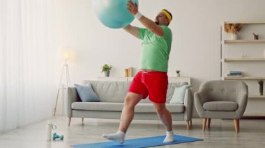 Tombul bir milenyum adamı evde antrenman yapar, fitness seansı için denge topu kullanır. Rahat bir spor kıyafeti giyer ve egzersizine odaklanır..