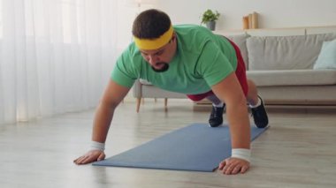 Yeşil tişörtlü ve kırmızı şortlu bir adam evinde mavi bir paspasın üzerinde çalışıyor. Kilo vermek için spor salonuna odaklanıyor. Güneşli bir iç mekanda azim ve çaba gösteriyor..