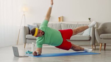 Bir milenyum adamı evde egzersiz yapıyor, yoga minderinde bir kalas yapıyor. Rahat spor kıyafetleri giyiyor ve rehberlik için online kaynak kullanıyor. Oturma odası aydınlık ve havadar..