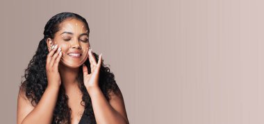 Bir kadın cilt bakımı rutininden zevk alıyor, cildine nazikçe yüz bakımı uyguluyor. Bu huzurlu ve izole ortamda rahatlık ve neşe saçarak güzelliğini ön plana çıkarıyor..