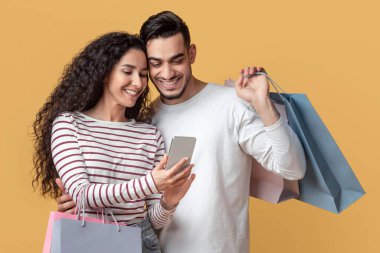 Alışveriş Uygulaması. Orta Doğu Eşleri Alışveriş Torbası Tutuyor ve Akıllı Telefon Kullanıyor, Satılık Teklif ve İndirimli Genç Arap Çift Tarama Uygulaması, Sarı Arkaplanda Duruyor