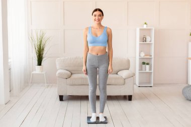 Kilo kaybı konsepti. Sıska kadın evde zayıfladıktan sonra tartıya çıkıp kameraya gülümsüyor. Boş Alan