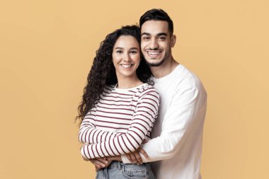 Kameraya sarılan ve gülümseyen genç Arap erkek ve kadının portresi, sarı arka planda birlikte poz verirken romantik Orta Doğu çifti,