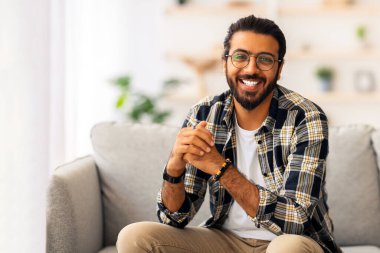 Yakışıklı orta doğu milenyumu adamı evde poz veriyor, günlük kıyafetler ve gözlükler takıyor, rahat oturma odasında koltukta oturuyor, kameraya gülümsüyor, hafta sonunu yalnız geçiriyor, boşluğu kopyalıyor.