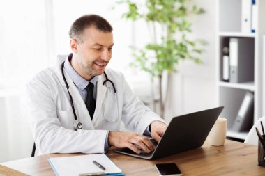 Araştırma Konsepti. Gülümseyen orta yaşlı bir uzman doktorun portresi bilgisayar bilgisayarı kullanıyor, klavyede daktilo kullanıyor, hastanede masada oturuyor, çevrimiçi konsolosluk veriyor, teletıp veriyor.