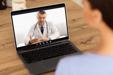 Teletıp Konsepti. Omzunun üstünden bakan kadın bilgisayarında doktorla çevrimiçi video görüşmesi yapıyor, evinde oturuyor. Uzaktan danışmanlık yapan profesyonel bir hekim.