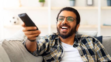 Evde televizyon seyreden, koltukta tek başına oturup mutlu bir yüz ifadesi takınan rahat bir Arap. TV kumandalı mutlu genç adam komedi izliyor ve gülüyor, kopyalama alanı, yakın çekim