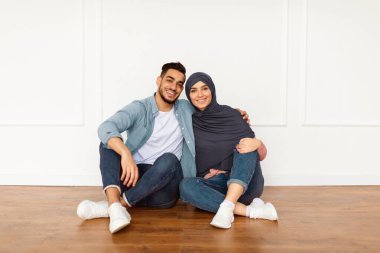 Sevgi dolu bir aile. Gülümseyen Müslüman kadın ve adamın portresi yeni dairede beyaz duvara yaslanmış şekilde yerde oturuyor. Mutlu çift kucaklaşıyor, kameraya bakıyor, taşınma gününü kutluyor.