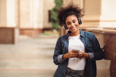 Cep telefonu kulaklığı takan mutlu Afrikalı öğrenci kameraya gülümsüyor ve kolej binasının yanında dikiliyor. Müzik ya da Eğitim Podcast 'i Dinleyen Genç Bayan