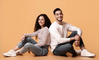 Sarı Stüdyo Arkasında Sırt Sırta Oturan neşeli genç Arap çift, Mutlu Milenyum Orta Doğulu Erkek ve Kadın 'ın Portresi Yerde Birlikte Rahatlıyor, Gülümsüyor ve Kamera,