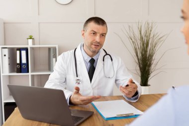 Reçeteyi açıklıyorum. Ciddi orta yaşlı bir doktorun portresi bilgisayarla masada oturuyor ve hastanedeki genç bir kadınla konuşuyor, modern klinikteki randevusu sırasında danışmanlık yapıyor.