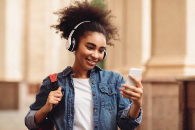 Cep telefonu mesajlaşması kullanan, interneti kolaçan eden, dışarıda üniversite binasının yanında duran kulaklık takan neşeli siyah öğrenci kız. Millennial Female Eğitim Podcast 'i Dinliyor