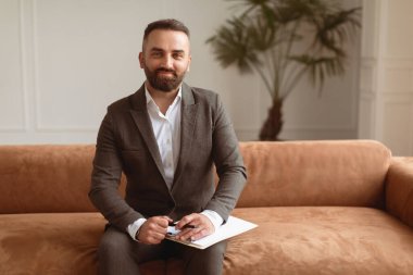 Confident Professional Therapist Concept. Portrait Of Happy Smiling Man Wearing Suit Sitting On Couch Sofa, Holding Clipboard With Pen And Posing Looking At Camera. Session At Office
