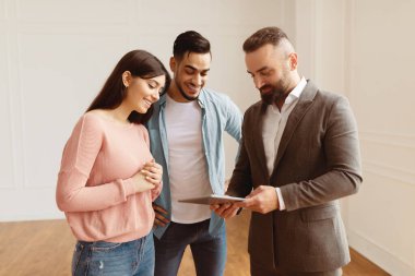 Professional Real Estate Agent In Suit Showing Digital Tablet With Build Project, Discussing House Plan, Selling Flat To Happy Family. Millennial Couple Buying New Apartment, Looking At Screen