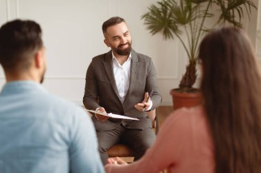 Aile Psikologu Konsepti. Ofiste oturan bir çiftle takım elbise giyen, elinde kalemle pano tutan, profesyonel tavsiyeler veren evli bir adamın portresi.