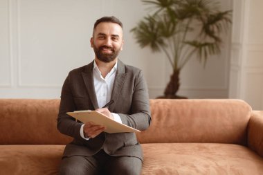Profesyonel Terapist Konsepti. Koltukta oturan gülümseyen adamın portresi, elinde pano, kalem, poz veren ve kameraya bakan. Ofiste Oturum