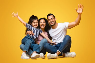 Sarı stüdyonun arka planında oturmuş eğlenen üç kişilik neşeli Arap ailesi, mutlu bir Orta Doğulu ebeveyn ve küçük kızları gülüyor ve ellerini kaldırıyor.
