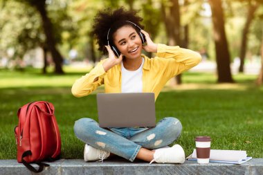 E-öğrenme. Dizüstü bilgisayar kullanan, kulaklık takan, konferans sonrası müzik dinleyen kampüs parkında oturan mutlu genç kız. Çevrimiçi Öğrenen Kadın Öğrenci
