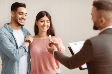 Satılık Emlak Konsepti. Mutlu gülümseyen çiftin portresi gelecekteki yeni dairenin anahtarını alıyor. Takım elbiseli profesyonel bir emlakçı genç alıcılara anahtar veriyor. İpotek