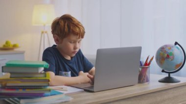 Kızıl saçlı bir çocuk evde çalışırken dikkatle dizüstü bilgisayarına odaklanıyor. Etrafı kitaplar ve bir küre ile çevrili, öğleden sonra online dersine dalmış..