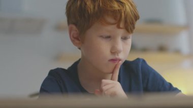 Kızıl saçlı genç bir çocuk evde çalışırken internet dersine dikkatle odaklanıyor. Derin düşüncelere dalmış, parmak kaldırmış, öğrenme sırasında konsantrasyonu gösteriyor..