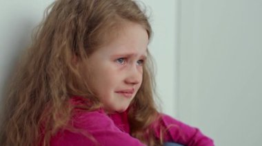 Kıvırcık saçlı genç bir kız duvara yaslanırken sıkıntı belirtileri gösterir. Yüzündeki ifade, aile içi şiddetin etkilerini gözler önüne seriyor. Zorlu bir ortamda hissedilen duyguları vurguluyor..