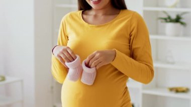 Müstakbel bir anne sarı uzun kollu bir gömlek giyer, kucağında bir çift pembe bebek ayakkabısı tutarken nazikçe şişlik yapar, evinde mutluluk ve beklenti saçar..