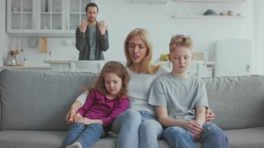 Bir anne iki çocuğuyla birlikte kanepede oturuyor, ikisi de sıkıntılı görünüyor. Arka planda, bir adam tartışıyor gibi görünüyor, evde gergin bir kargaşa yaratıyor..