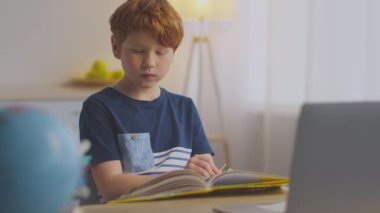 Kızıl saçlı reşit olmayan bir çocuk bir masada oturup online öğrenimine konsantre oluyor. Önünde açık bir kitap ve iyi aydınlatılmış bir alanda yanında bir laptop var..