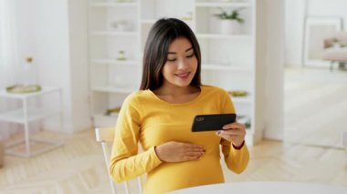 Asyalı hamile kadın karnını tutarken gülümser ve parlak, karşılama odasında akıllı telefonuna bakar. Hamileliğiyle ilgili içeriklerle meşgul gibi görünüyor..