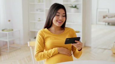 Sarı uzun kollu tişörtlü Asyalı bir kadın telefonuna gülümserken bebek şişliğini kucağına alır. Evinde rahat bir şekilde oturuyor. Etrafı aydınlık ve neşeli bir çevreyle çevrili..