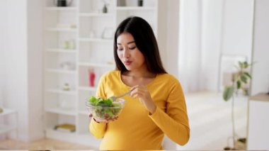 Sarı bluzlu Asyalı hamile kadın parlak ve serin oturma odasında dururken bir kaseden taze salata yiyor. Memnun görünüyor ve sağlıklı yemek seçimine odaklanmış..