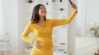Mutlu bir Asyalı hamile kadın, kapalı bir ortamda selfie çekerken poz veriyor. Bu özel anın tadını çıkarırken eli karnına hafifçe dayanıyor..