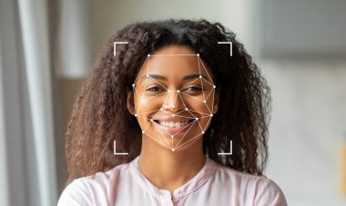 Kıvırcık saçlı, parlak gülümseyen, yüz hatlarını gösteren bir kadın. Üstü kapalı grafikler yüz tanıma teknolojisinin kullanıldığını gösteriyor. Ortam aydınlık ve davetkar..