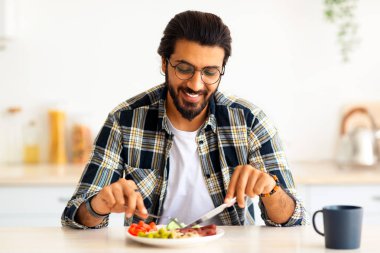 Olumlu Arap adam hipster evde sağlıklı kahvaltı yapıyor, taze sebze yiyor, sosis yiyor, kahve içiyor, mutfak masasında oturuyor, fotokopi çekiyor. Yakışıklı Hintli adam güne güzel yemeklerle başlıyor.