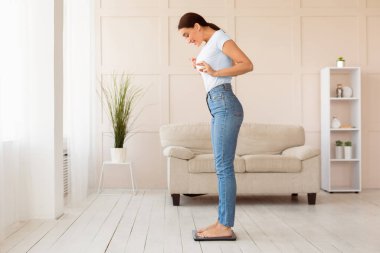 Kilo kaybı konsepti. Kilo tartısı üzerinde duran kız evde zayıf parmaklarını çapraz yaparak tartıyor. Boşluğu Kopyala