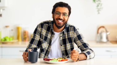 Keyifli, orta doğulu, sakallı, günlük kıyafetleri ve şık bardakları olan, evde sağlıklı bir öğle yemeğinin tadını çıkaran, mutfak masasında oturan, sebze salatası yiyen, çay içen, sağlıklı deit kavramı olan genç bir adam.