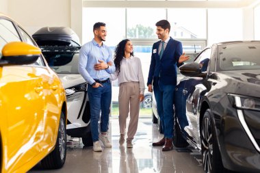 Yakışıklı Arap satış elemanı genç aile babasını ve karısını en yeni araba galerisine veriyor, neşeli satıcı mutlu orta doğu çifti için uygun siyah otomobil gösteriyor, tam boy şut.