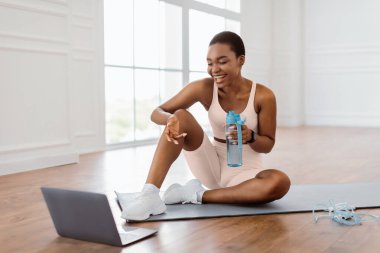 Çevrimiçi ev eğitimi. Özel antrenör ya da takipçileriyle webcam 'e konuşan, yoga minderinin üzerinde dizüstü bilgisayarın önünde oturan mutlu Afrikalı kadın. PC ya da yayında bayan kayıt öğreticisi