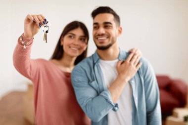 Pozitif genç çift evin anahtarını gösteriyor, yeni evin oturma odasında duruyor, seçici odak eli. Milenyum Arap erkeği ve bayanı daha büyük bir daireye taşınmaktan memnuniyet duyar, ellerinde düz anahtar, bulanık arka plan