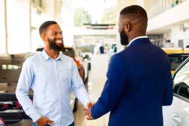 Araba almak. Başarılı bir anlaşmadan sonra galeride el sıkışan siyah satıcı ve erkek müşterinin portresi, modern oto galerisinde yeni otomobil satın alan gülümseyen Afrikalı Amerikalı adam,