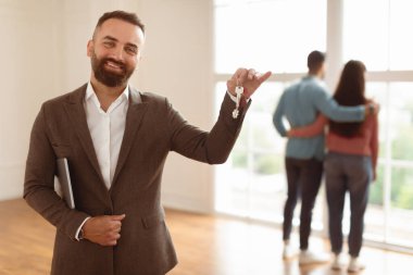 House Ownership. Portrait Of Smiling Professional Real Estate Agent In Suit Holding And Showing Key To Camera, Selective Focus. Happy Spouses Choosing New Home Looking Out Window In Blurred Background