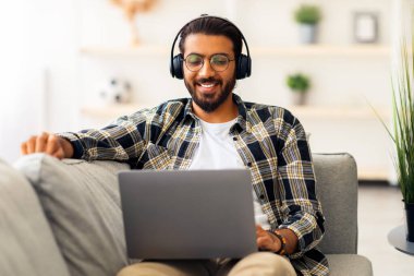 Uzun saçlı, sakallı, orta doğulu adam internetten film izliyor, kanepeye yaslanıyor ve oturma odasında dizüstü bilgisayar ve kulaklık kullanıyor. Notebook üzerinden görüntülü arama yapan pozitif Arap adam, boşluğu kopyala