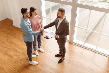 Başarılı bir anlaşma, gayrimenkul, ev satın alma sözleşmesi. Mutlu gülümseyen milenyum alıcıları yeni dairede profesyonel emlakçıyla el sıkışıyor. Birlikte bir anlaşma yapıyorlar.