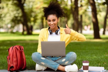 E-öğrenme. Afro-Amerikalı öğrenci dizüstü bilgisayar kullanıyor ve üniversitenin kampüs parkında açık havada oturmak için eğitici web sitesini onaylıyor. Uzak Eğitim