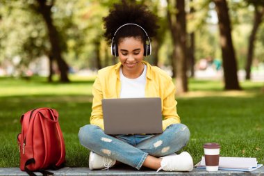 E-öğrenme. Diz üstü bilgisayar kullanan, kulaklık takan, dışarıdaki parkta battaniyenin üzerinde oturan siyah bir kız öğrenci. Teknoloji ve Öğrencilik. Modern Uzaktan Eğitim Konsepti.